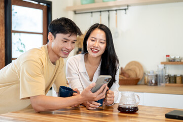 Obraz premium Smiling asian woman and man couple looking at phone watching videos making coffee at kitchen counter