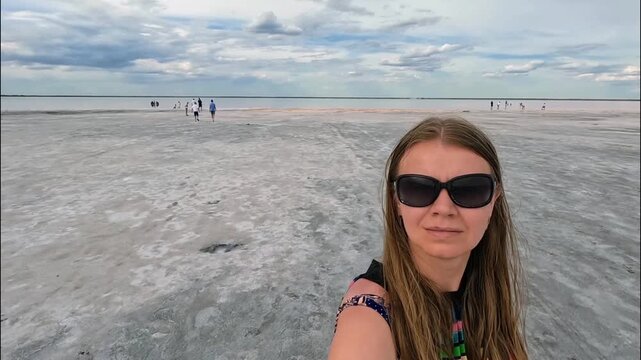 A woman in sunglasses takes a selfie on a salt marsh by the sea. In the distance, people are walking along the shallows. Travel, summer, spaciousness, copyspace.