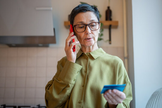 Concerned elder woman talking on smartphone with scammers, holding credit card and sharing personal data to fake bank employees. Vulnerable mature female tricked by financial manipulators by phone.