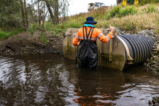 Worker in hi-vis safety gear and waders installs large flood control gate in stream with culvert pipe. Perfect for environmental engineering, water management companies, and infrastructure development
