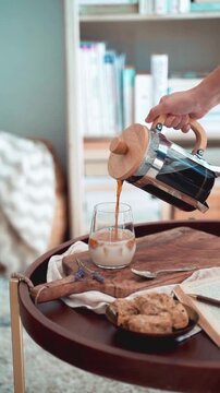 Pouring fresh coffee from French press into a glass of milk on a wooden tray