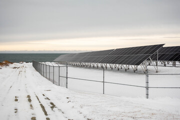 Solar panels mounted on metal structures stand on a snowy meadow beside the ocean in Canada. Clean energy technology placed in a cold coastal environment with sea views.