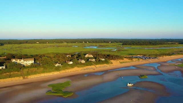 Drone aerial over Cape Cod barrier beach, sandy bluff and coastal homes beside a busy beach access area, with braided marsh creeks and wetlands beyond. Travel lifestyle b-roll, cinematic 4K.