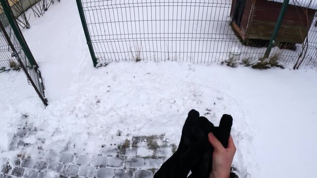 POV removing a black winter glove in a snowy backyard near a wire fence and icy paving stones, pausing outdoor work during cold weather home chores.