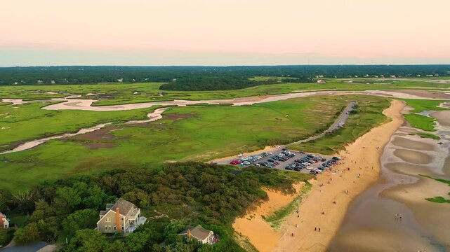 Drone aerial over Cape Cod beach and dunes at low tide, revealing a packed parking lot by the shore, winding salt marsh channels inland, and wide sandy flats. Coastal summer 4K b-roll.