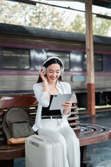 Smiling woman with headphones and tablet at a train station, ready for travel with suitcase and...