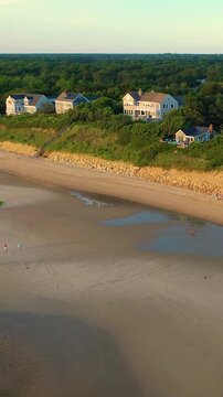 Vertical drone golden hour over Cape Cod beach and dunes, coastal homes along the shore with marshland stretching inland. Tall-frame view of low tide sand and warm light, cinematic b-roll.
