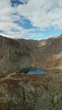 Raw drone footage executes a high-altitude reverse dolly-out shot in the mountains, revealing an autumn mountain lake below. The frame captures the full spectrum of colors from the mountain flora duri