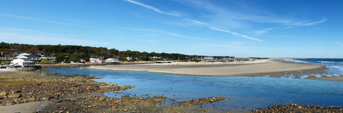 Panoramic view of Ogunquit River tidal estuary opening toward Atlantic shoreline, expansive sand flats, shallow blue channel, rocky foreground, seaside residences