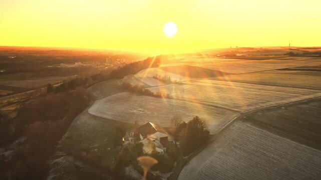 Forward drone flight near Elchingen during golden-hour sunset, gliding over rolling farmland and frosty fields with warm sun flares and long shadows.
