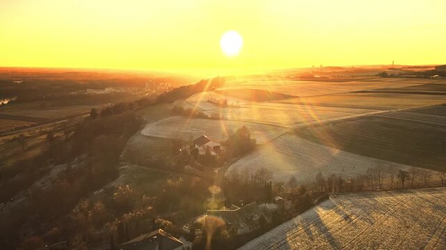 Forward drone flight near Elchingen during golden-hour sunset, gliding over rolling farmland and frosty fields with warm sun flares and long shadows.