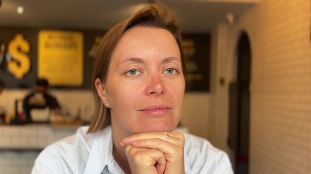 A woman sits in a cafe with her chin resting on her hands. She is lost in thought while enjoying her coffee. The background shows other customers and a counter.