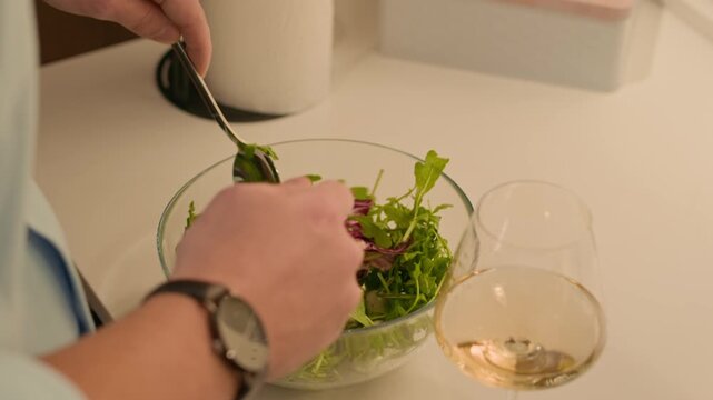 Close up shot of hands of unrecognizable man mixing green salad in bowl at kitchen table with white wine glass during home dinner