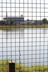 Obraz premium View of industrial buildings across a calm lake seen through a wire fence