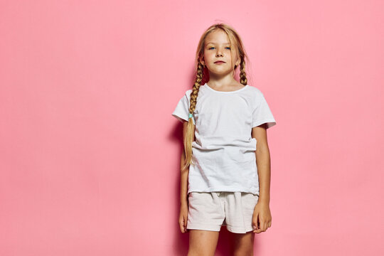 Young girl with long braided hair stands confidently in white t-shirt and shorts against pink background. Studio portrait of child.