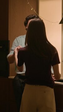 Vertical shot of affectionate Caucasian boyfriend and girlfriend slow dancing under warm lighting at home