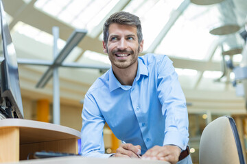Happy businessman taking notes while working in modern office, standing by his desk, using a modern...