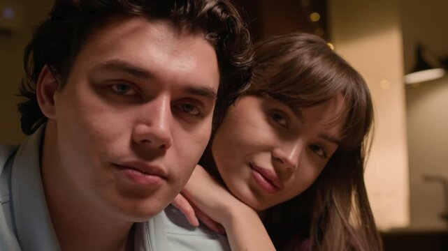 Close up portrait of cheerful Caucasian young boyfriend and girlfriend looking at camera while sitting under warm home lighting