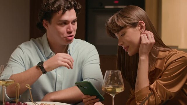 Medium handheld shot of young Caucasian couple having discussion using smartphone while dining with homemade pasta and white wine