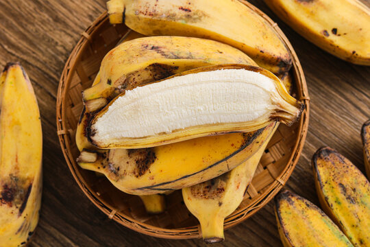 Top view of a woven basket filled with ripe yellow saba bananas (pisang kepok). With one fruit peeled to show its center. The wooden surface creates a warm and organic scene.