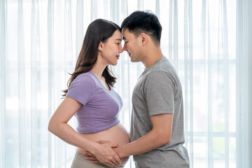 Fototapeta premium Asian young pregnant woman gently stroke belly with husband in house.