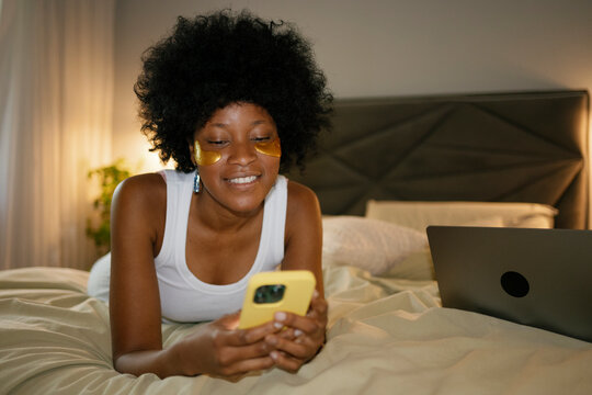 Woman laughing and using smartphone with eye patches in bed in the morning