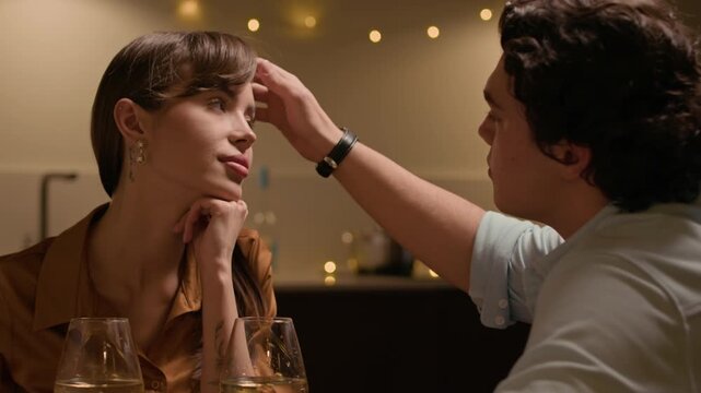 Medium close up shot of gentle Caucasian man brushing his girlfriends hair back having intimate moment during romantic dinner at home