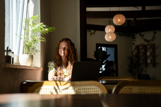 Woman with red hair working remotely in cafe near window with laptop