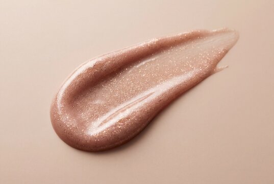 Smear of creamy rose gold lip gloss with shiny glitter particles on beige surface