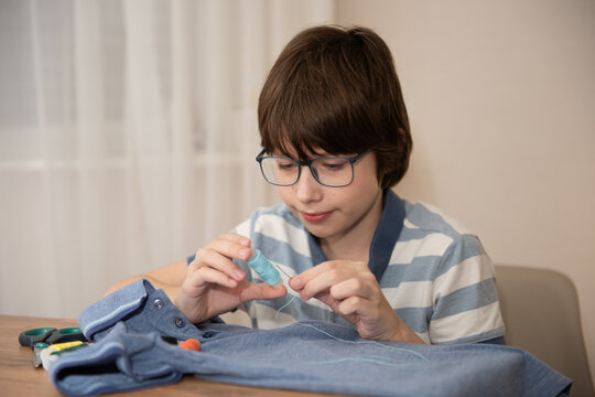 Young boy sewing a button on a denim shirt at a home table, focused on stitching and mending the garment.