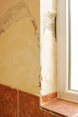 A damaged corner wall with falling plaster next to a window