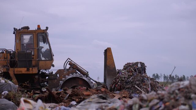 Heavy industrial machinery working at a large landfill site, with a special bulldozer compacting and managing piles of garbage and solid waste against a gray, overcast sky