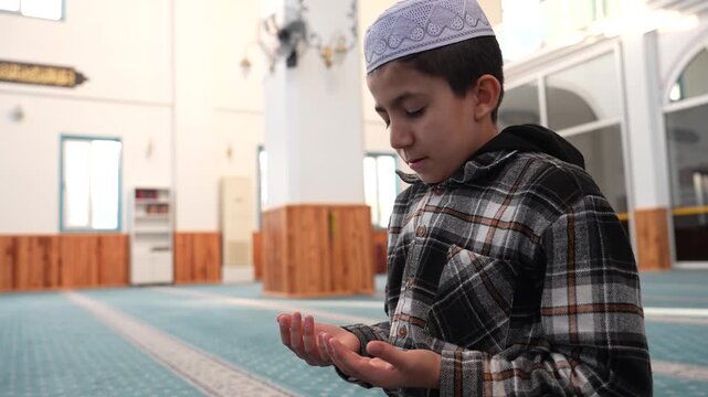 Muslim boy making Dua and praying in a mosque with devotion. Young child supplicating during Ramadan at masjid. Muslim kid praying with hands raised in a spiritual moment.