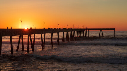 Fototapeta premium The Pacifica Municipal Pier Silhouetted Against Glowing Sunset in Orange and Gold. Pacifica, San Mateo County, California.