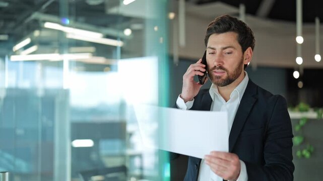 Confident business professional converses on phone, reviewing documents in contemporary office. Serious demeanor reflects dedicated work ethic, strategic communication.