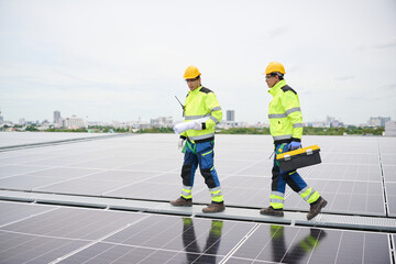 Two construction workers walking on a rooftop solar panel array, one carrying a blueprint, another with a toolbox. They wear safety gear, indicating a professional environment