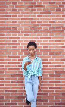 Smiling woman leaning against brick wall using earphones and cell phone