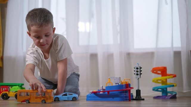 Preschooler in socks drives toy bus forward. Boy smiling while arranging vehicles. Toddler enjoys playful moment, young learner interacting. Child engages in role-play with school bus on floor.