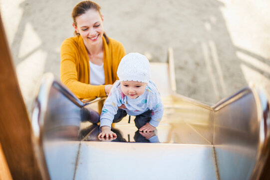 Baby girl climbing on shute on playground being held by her mother
