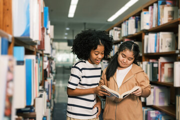 Two young students smile while reading a book together between bookshelves in a well-lit library.