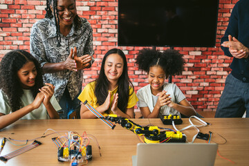 A diverse group of kids and an adult enthusiastically celebrate a successful robotics experiment together at a table in a creative indoor setting. © artitwpd