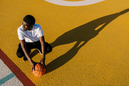Thoughtful young man holding basketball crouching on sports court during sunny day