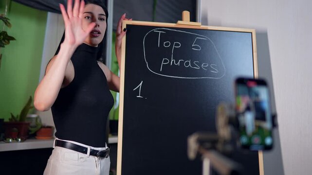 Uncertain actress is trying to film a video on smartphone standing next to the blackboard but forgetting words and failing starting to laugh. Caucasian woman is streaming a lesson explaining material.