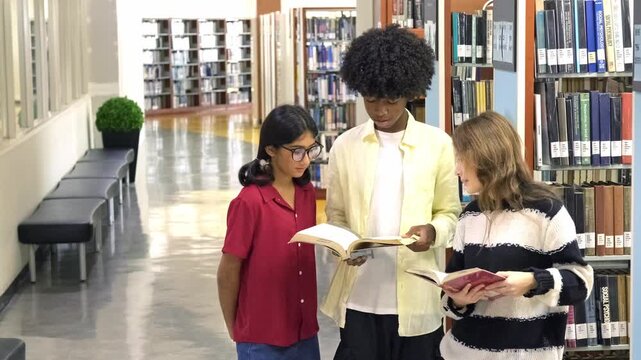 Three cheerful students stand in a bright library hallway, smiling and comparing books beside tall shelves, symbolizing youth education, learning resources, and academic engagement.