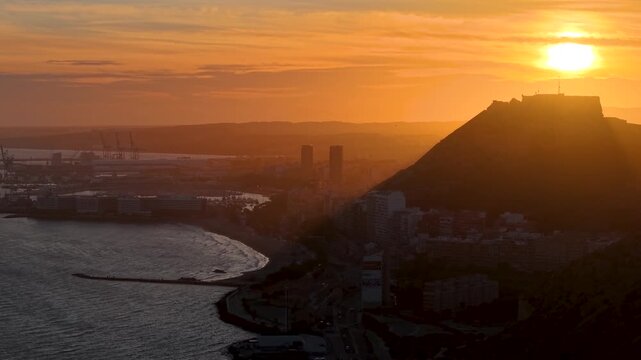 Alicante al atardecer desde el mar con el sol detr&aacute;s del Castillo de Santa Barbara , panor&aacute;mica a&eacute;rea