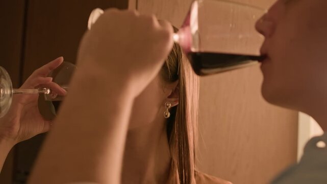 Close up shot of affectionate young Caucasian couple clinking glasses with red wine and communicating while having date at home in warm light
