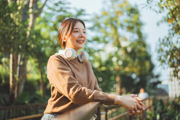 Happy Asian woman leaning on balcony railing in green park wearing sweater and headphones. Outdoor lifestyle relaxation and mental wellness concept with natural sunlight. Enjoying morning in city