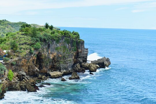 A beautiful view of Ngobaran Beach in Yogyakarta, Indonesia. Blue sea with blue sky. Waves crashing against the rocky cliffs. A view of the green rocky cliffs and the sea from above.