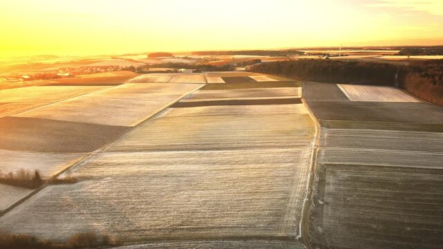 Forward drone flight over a patchwork of frosty agricultural fields at golden hour, with warm sunrise glow, gentle haze, and distant countryside on the horizon.