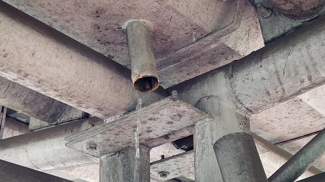 Water leaking from metal pipe under concrete structure showing infrastructure damage, maintenance issue, urban drainage problem, construction defect and water wastage concept